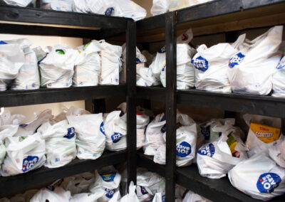 The camera points at where two shelves laden with bags of food meet. The bags are made of white plastic and are overflowing with boxed and canned foods for families to come pick up and take home.