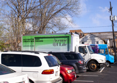 Parked next to a line of cars in a parking lot sits a medium-sized box truck. The cab of the truck is white but the sides of the cargo hold are bright green at the top before fading to white at the bottom. The top right side of the cargo hold has the Avenue logo depicted on top of a photo of a stack of mattresses. The top left of the cargo hold has black text reading “OLD MATTRESSES CREATE NEW DREAMS” with Avenue of Life’s website written in black text below.