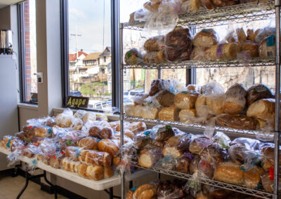 A five-tiered metal utility shelf stands next to a large folding table. Both are overflowing with dozens of bagged bread products that vary in type.
